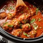 a crockpot with meatballs in tomato sauce with a wooden spoon