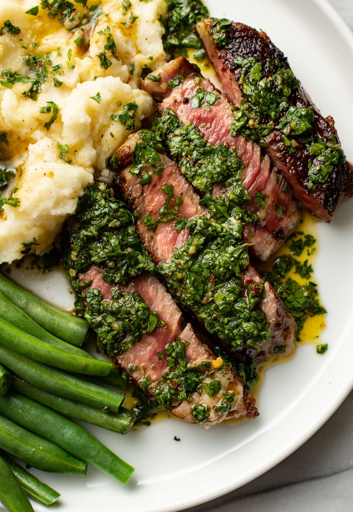 a plate with ribeye steak, chimichurri, green beans, and mashed potatoes